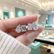 A close-up of a hand holding a pair of stud earrings with pear, baguette, and round-cut clear gemstones, set against a softly blurred jewelry store background with turquoise accents.
