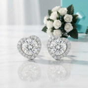 A pair of heart-shaped stud earrings with large round clear center stones surrounded by smaller halo stones, displayed on a light reflective surface with white roses and a turquoise box in the background.