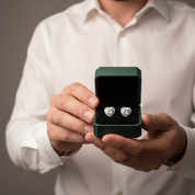 A person in a white dress shirt holds an open dark green jewelry box displaying a pair of silver heart-shaped stud earrings with clear gemstones.