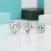 A pair of silver heart-shaped stud earrings with clear gemstones, shown on a reflective white surface with a blurred turquoise and white background.