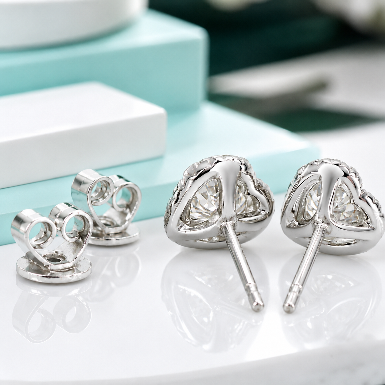 Silver heart-shaped stud earrings with clear gemstones, shown from both front and back views on a reflective white surface with light blue jewelry boxes in the background.