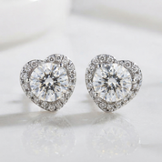 A pair of heart-shaped stud earrings with large clear center stones surrounded by a halo of small clear stones, set in a silver-toned metal on a light reflective surface.