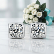 A pair of square stud earrings with large round clear gemstones in a silver-toned bezel setting, displayed on a light marble surface with white flowers and a turquoise ribbon in the background.
