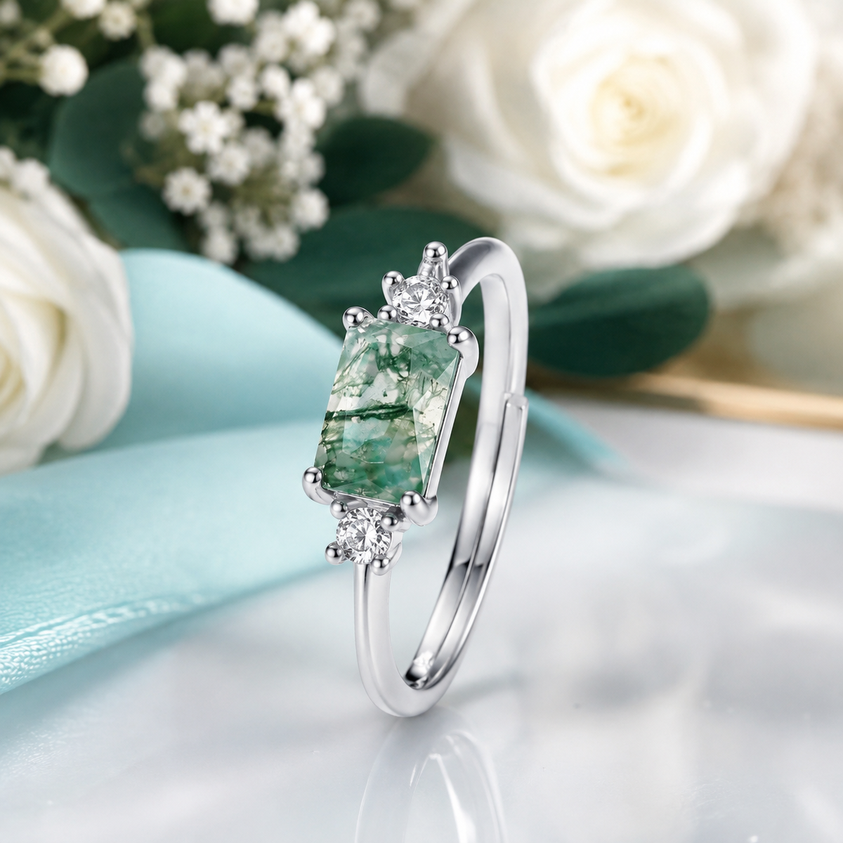 A silver ring with a rectangular green emerald-cut center stone and small clear accent stones, displayed on a white marble surface with white roses and a light blue ribbon in the background.