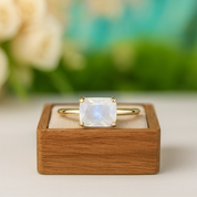 Gold ring with a square-cut gemstone on a wooden box against a blurred floral background