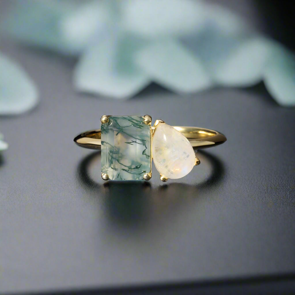 Gold ring with a square green gemstone and a teardrop white gemstone on a dark surface