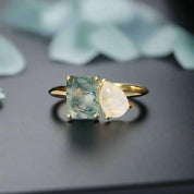 Gold ring with a square green gemstone and a teardrop white gemstone on a dark surface
