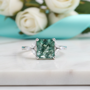 Green gemstone ring with diamond accents on a white surface with flowers in the background