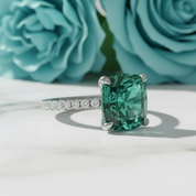 Green gemstone ring with diamond accents on a white surface with teal roses in the background