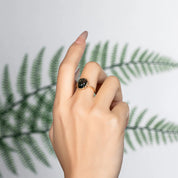 Hand wearing a gold ring with a dark gemstone against a light background with green leaf shadows