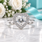 A heart-shaped diamond ring with a halo of small diamonds on a silver band, displayed on a white marble surface with white roses, baby's breath, and a turquoise ribbon in the background.