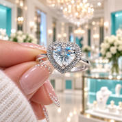 A heart-shaped diamond ring with a halo setting held by a hand with manicured nails, set against a blurred luxury jewelry store background.