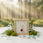 Jewelry box with a necklace featuring a red gemstone, surrounded by flowers and moss in a forest setting.