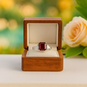 Jewelry box with a ring featuring a red gemstone, set against a blurred floral background.