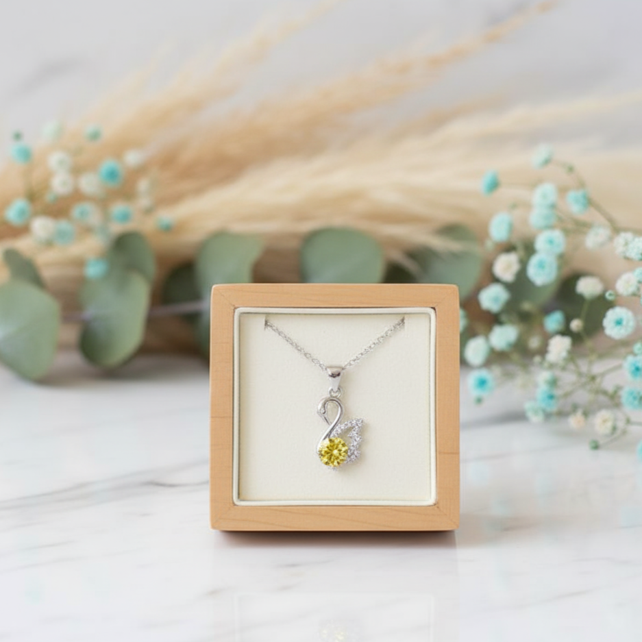 Necklace with a yellow heart pendant in a wooden box, surrounded by flowers and greenery.