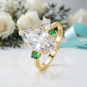 A gold ring with a large marquise-cut clear center stone and two small green emerald-cut side stones, displayed on a white surface with blurred white roses and a light blue ribbon in the background.
