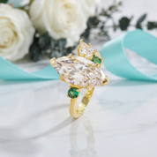 A gold ring featuring a large marquise-cut clear gemstone and smaller round and oval green gemstones, displayed on a light marble surface with white roses and a turquoise ribbon in the background.
