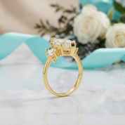 A yellow gold ring with a three-stone cluster setting featuring clear gemstones, displayed on a white marble surface with a blurred background of white roses and a turquoise ribbon.
