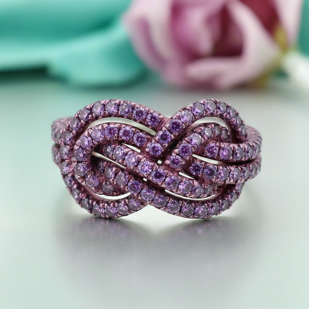 Purple knot-shaped ring on a light green background with a blurred pink flower in the background
