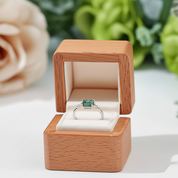 Ring with green gemstone in a wooden box on a white surface with flowers in the background