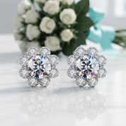 A pair of silver-toned flower-shaped stud earrings with clear sparkling gemstones, displayed on a white reflective surface with a blurred bouquet of white roses and a turquoise ribbon in the background.