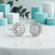 A pair of silver-toned flower-shaped stud earrings with clear gemstones, displayed on a white marble surface with a soft-focus background of turquoise and white geometric shapes.