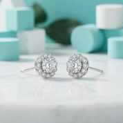 A pair of round diamond halo stud earrings in silver-toned metal, displayed on a white marble surface with a soft-focus turquoise and white background.