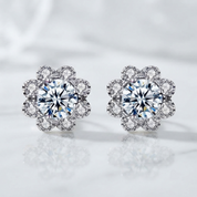 A pair of silver-toned flower-shaped stud earrings with large round clear center stones and smaller surrounding stones, shown on a soft white reflective surface.