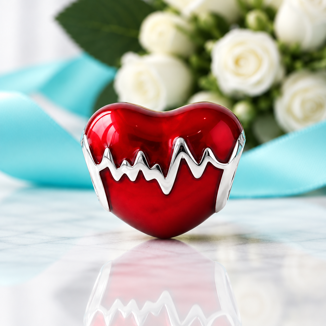 A glossy red heart-shaped pendant with a silver EKG line detail, displayed on a reflective marble surface with a blurred bouquet of white roses and a light blue ribbon in the background.