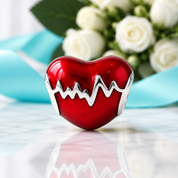A glossy red heart-shaped pendant with a silver EKG line detail, displayed on a reflective marble surface with a blurred bouquet of white roses and a light blue ribbon in the background.