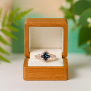 Rose gold ring with a blue gemstone in a wooden box against a blurred green background