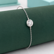 A delicate silver chain bracelet with a single bezel-set round clear gemstone, displayed on a dark green velvet jewelry roll.