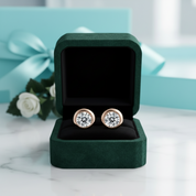 A pair of round stud earrings with clear gemstones in a gold-tone setting, displayed inside an open dark green jewelry box on a white marble surface, with a blurred turquoise gift box and white rose in the background.