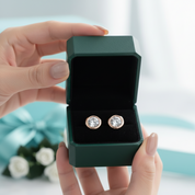 A pair of round stud earrings with clear gemstones in gold-toned settings, displayed inside an open dark green jewelry box held by two hands.