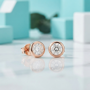 A pair of round rose gold stud earrings with clear gemstones, displayed on a reflective white surface with a soft-focus turquoise and white background.