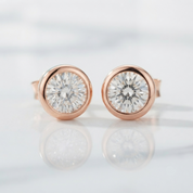 A pair of round stud earrings with brilliant-cut clear gemstones in rose gold bezel settings, displayed on a reflective white marble surface.