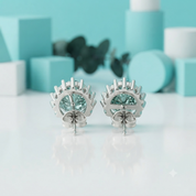 A pair of silver-toned earrings with large round light blue gemstones, displayed on a reflective white surface with a soft-focus turquoise and white geometric background.