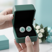 A pair of round stud earrings with a light blue-green center and silver-toned floral halo settings, displayed inside an open dark green jewelry box held by hands.