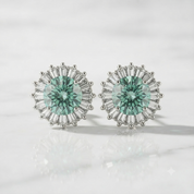 A pair of silver-toned stud earrings featuring round mint green center stones surrounded by baguette and round clear accent stones, displayed on a light marble surface.