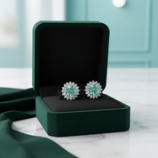 A pair of round stud earrings with light blue and white floral designs, displayed inside an open dark green jewelry box on a white marble surface.