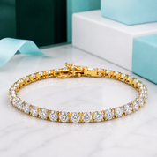 A gold tennis bracelet set with round-cut clear stones, displayed on a white marble surface with light blue gift boxes in the background.