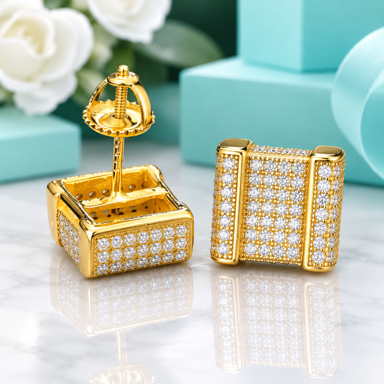 A pair of gold square stud earrings covered in small clear stones, shown on a polished marble surface with turquoise blocks and white flowers in the background. One earring is front-facing and the other is turned to show the back post.