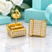 A pair of gold square stud earrings covered in small clear stones, shown on a polished marble surface with turquoise blocks and white flowers in the background. One earring is front-facing and the other is turned to show the back post.