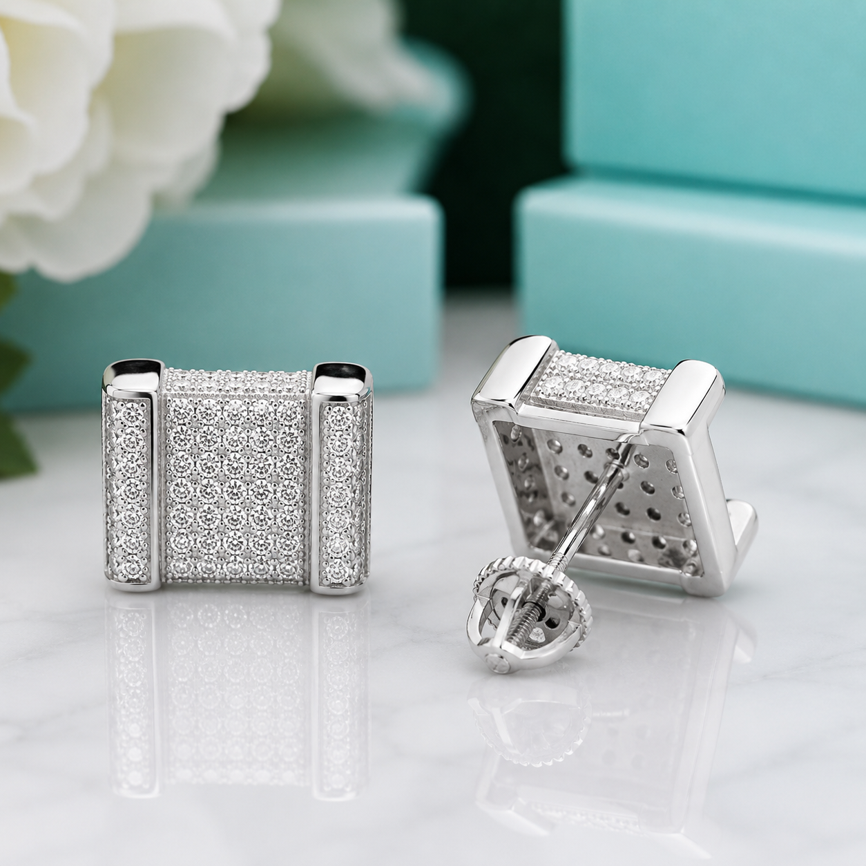 A pair of silver-toned square stud earrings covered in small round clear stones, shown on a white marble surface with a turquoise box and white flower in the background.