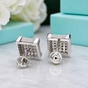A pair of silver-toned cube stud earrings with pavé-set clear stones on the front and a textured geometric pattern on the back, displayed on a white marble surface with blurred turquoise gift boxes and white flowers in the background.