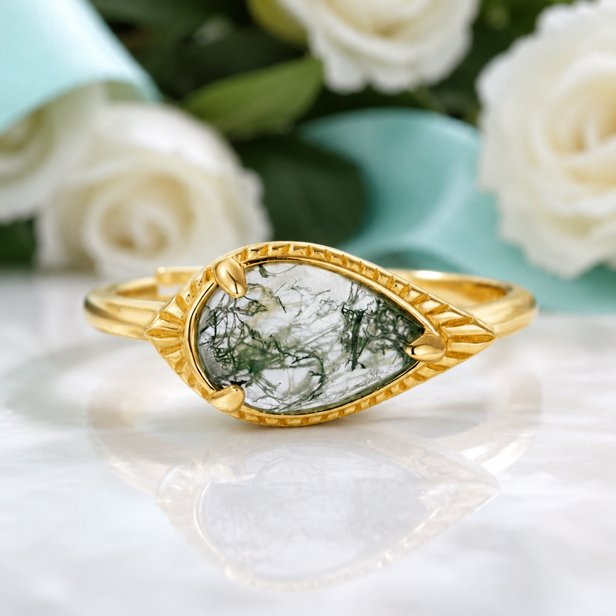 A gold ring with a marquise-shaped clear gemstone containing green and white crystalline inclusions, displayed on a white marble surface with blurred white roses and a light blue vase in the background.