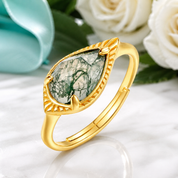 A gold ring with a teardrop-shaped green and white marbled stone, styled on a white marble surface with white roses and a turquoise flower in the background.