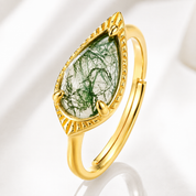 A gold ring featuring a teardrop-shaped clear stone inlaid with green tree-like fibers, set in a textured gold bezel on a white background.