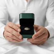 A gold ring with a marquise-cut light blue gemstone is displayed in an open dark green jewelry box held by a person wearing a white button-down shirt.