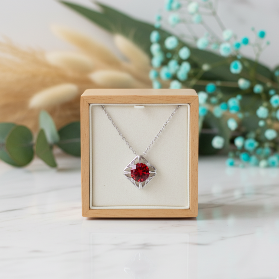 Silver necklace with a red gemstone in a wooden box on a light surface with decorative elements.